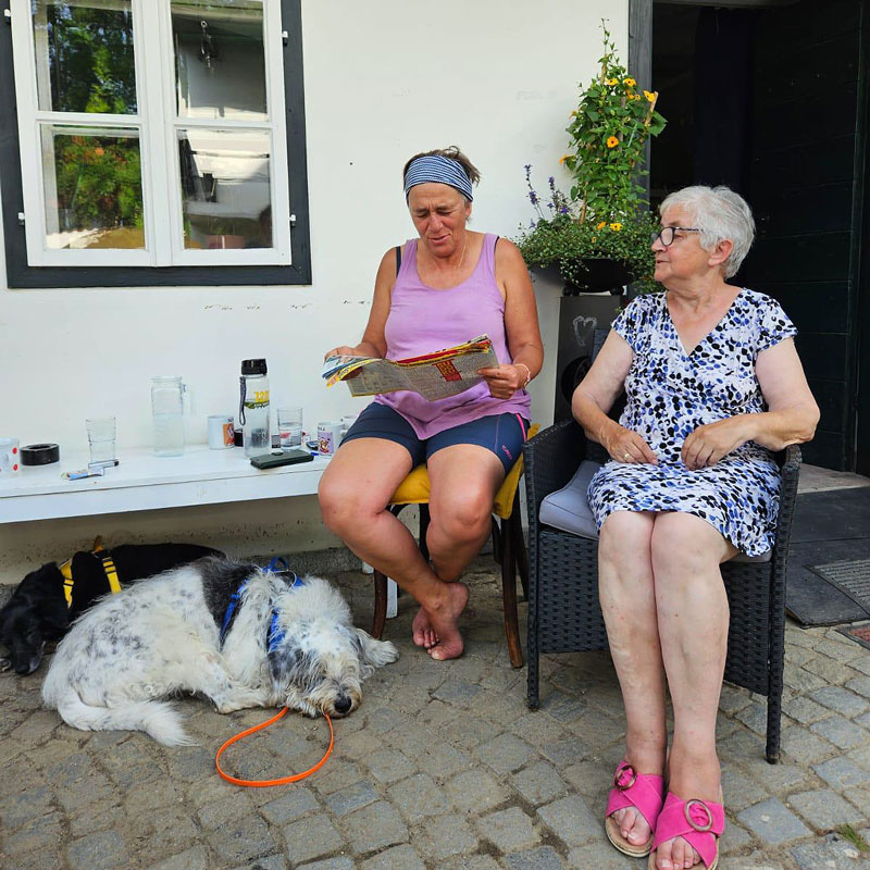 Bild von zwei älteren Damen die gemütlich auf einem Gartenstuhl sitzen. Zu ihren Füßen liegt ein großer, weißer Hunde